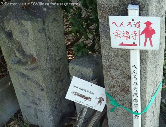 This is a Shikoku Pilgrimage (四国遍路 / Shikoku Henro) route marker pointing the way to Eifuku‑ji (栄福寺), Temple #57 on the 88‑temple pilgrimage; the smaller board is a local cemetery or community notice to direct visitors to nearby memorial sites, 2026-04-05, Imabari Castle, P. Potter.